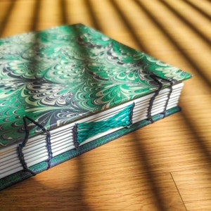 Könnte beinhalten: Ein handgefertigtes Notizbuch mit marmoriertem grünem, schwarzen und weißen Einband. Die Seiten sind mit schwarzem Faden und einem türkisfarbenen Akzent gebunden. Das Notizbuch liegt auf einer Holzoberfläche.
