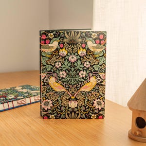 Puede incluir: Un diario de tapa dura rectangular con fondo negro y un patrón repetitivo de pájaros amarillos, flores rosas, follaje verde y bayas rojas. El lomo está encuadernado con costuras de hilo rosa visibles a lo largo del borde izquierdo.