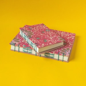 Pode incluir: Dois diários feitos à mão com capas estampadas florais em tons de vermelho, castanho e verde-azulado. As páginas são encadernadas com fio verde-azulado, e os diários são empilhados sobre um fundo amarelo. Artigos de papelaria únicos.