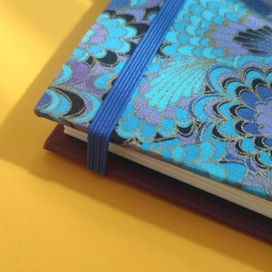 Puede incluir: Primer plano de un cuaderno con una cubierta de patrón floral azul y un cierre de banda elástica azul. La cubierta presenta tonos de azul, morado y dorado. El fondo es amarillo liso.