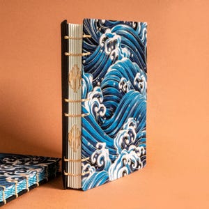 Puede incluir: Un diario hecho a mano con una cubierta con un diseño de olas azules y blancas. La columna vertebral es negra con costuras visibles e hilo dorado. Un artículo artístico único.