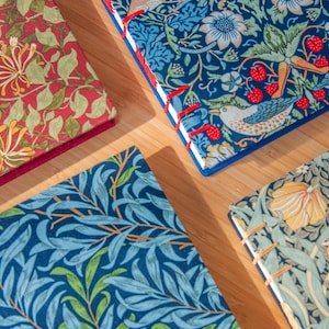 Op de afbeelding: Vier rechthoekige notitieboeken met stoffen kaften met ingewikkelde bloemen- en vogelpatronen. De notitieboeken tonen rode, blauwe en groene kleurenpaletten met zichtbare stiksels in rood en beige garen langs de randen van de rug.