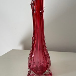 May include: A tall, slender, red glass vase with a textured, tree-like design. The vase has a wide base with a clear glass finish.