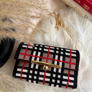 May include: A rectangular handbag featuring a black, white, red, and tan plaid pattern. The bag is constructed with a grid-like texture and includes a gold-toned metal bar clasp closure on the front flap, along with a matching gold-toned chain strap.