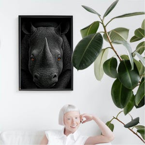 Könnte beinhalten: Ein gerahmtes Schwarz-Weiß-Porträt eines Nashorns. Die strukturierte Haut und das markante Horn des Nashorns sind detailliert. Die Augen des Nashorns sind von auffälliger orange Farbe. Das Kunstwerk wird an einer weißen Wand ausgestellt.