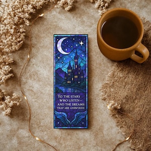 May include: A stained glass bookmark with a blue, purple, and black color scheme. The bookmark features a castle with a crescent moon and stars in the night sky. The text reads "To the stars who listen and the dreams that are answered".