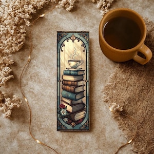 May include: A bookmark featuring a stack of books with a teacup on top, designed in a stained-glass style. The bookmark has a teal border and floral accents. A mug of coffee sits beside the bookmark on a neutral surface.