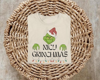 Può includere: Una maglietta color crema con un personaggio verde del Grinch che indossa un cappello rosso da Babbo Natale. Il testo "NICU GRINCHMAS" è stampato in nero sotto il personaggio. Una serie di luci colorate è stampata sotto il testo.