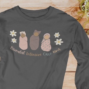 Pu&ograve; includere: Una maglietta a maniche lunghe grigio scuro con una grafica di tre neonati avvolti in coperte. I neonati sono circondati da fiori bianchi e dal testo "Neonatal Intensive Care Unit".
