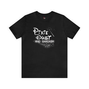 May include: Black t-shirt featuring a white graphic that reads "Pixie Dust and Sarcasm" in a stylised font. The graphic also includes a white wand with sparkles.
