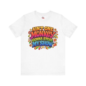 May include: White t-shirt with a colorful retro design. The graphic reads "Ain't One Monkey Gonna Steal My Show" in a vibrant, multi-colored font with stars and flower accents. The shirt is made of a soft material.