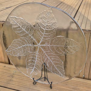 May include: A clear, round glass plate with a detailed leaf design etched into the surface. The plate is supported by a black metal stand, showcasing the intricate veining of the leaves. The plate is a decorative piece.