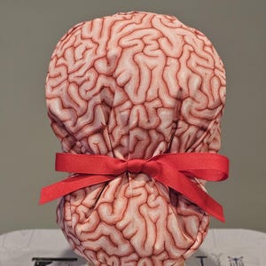 May include: A pink and red surgical scrub hat with a brain pattern. The hat is tied with a red ribbon.