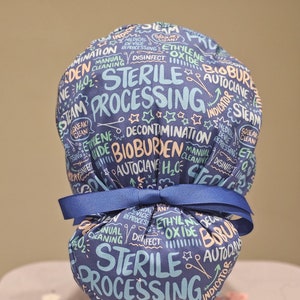 May include: A blue scrub hat with a white ribbon. The hat is covered in colourful text, including "Sterile Processing", "Bioburden", "Autoclave", and "Decontamination".