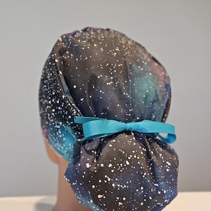 Op de afbeelding: Een zwarte scrub-muts met een galaxy print en witte stippen. De muts heeft een turquoise lint dat aan de achterkant in een strik is gebonden.