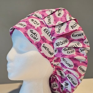 Può includere: Un berretto chirurgico rosa con bolle di dialogo bianche stampate con termini medici come "Bisturi", "Adson", "Pinza per asciugamano", "Aspirazione" e "Kelly".