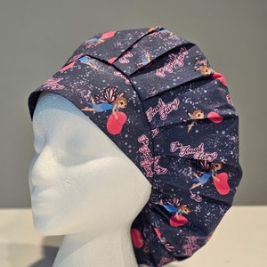 Pode incluir: Um gorro de cirurgia azul com um design plissado. O gorro apresenta um padrão repetido de fadas cor-de-rosa com asas e o texto "The Tonsil Fairy".
