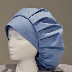 May include: A light blue bouffant surgical scrub cap. The cap is made of a soft fabric and features a gathered elastic back for a secure fit. The front of the cap has a pleated design. The cap is displayed on a white mannequin head.