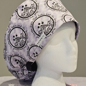Op de afbeelding: Een paarse en witte scrubmuts met een herhalend patroon van sterren en een geometrisch ontwerp. De tekst "To the stars that listen and the dreams that are answered" wordt herhaald op de muts.