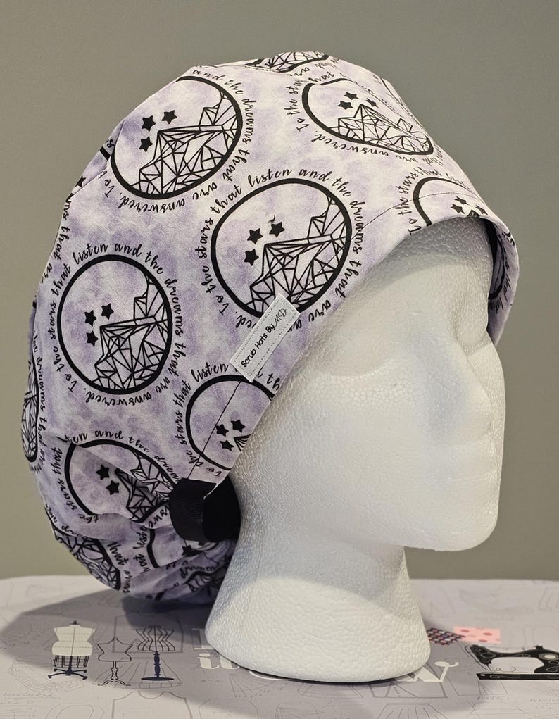 Op de afbeelding: Een paarse en witte scrubmuts met een herhalend patroon van sterren en een geometrisch ontwerp. De tekst "To the stars that listen and the dreams that are answered" wordt herhaald op de muts.