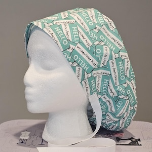 Pode incluir: Uma touca cirúrgica azul-turquesa e branca com um padrão repetido de texto "HELLO" e "Hey Anesthesia". A touca tem uma fita branca para amarrar e é exibida em uma cabeça de manequim branca.