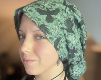 May include: A scrub cap featuring a light green background with a repeating pattern of black crows in flight, intertwined with dark green leafy vines and small white flowers. The fabric head covering is designed to fit securely around the head.
