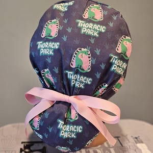 May include: A blue scrub hat with a pink tie. The hat has a repeating pattern of a green and pink dinosaur with the text "Thoracic Park".