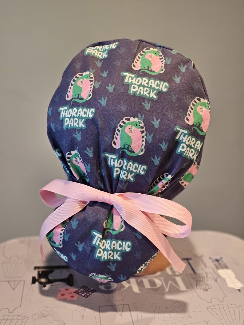 May include: A blue scrub hat with a pink tie. The hat has a repeating pattern of a green and pink dinosaur with the text "Thoracic Park".