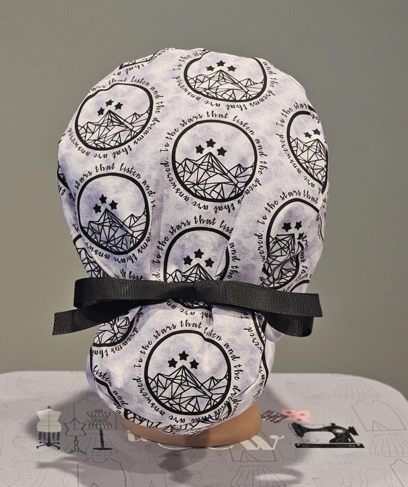Op de afbeelding: Een witte scrubmuts met een zwart-witte print van een bergketen en sterren. De tekst "Ik heb gedroomd van de sterren die luisteren en de dromen die worden beantwoord" wordt herhaald rond de muts.