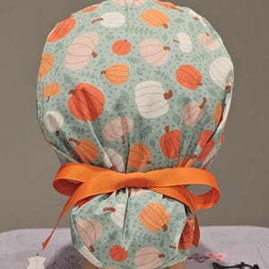 May include: A green scrub hat with a pumpkin print and an orange ribbon tie. The hat is made of fabric and has a white background with orange and white pumpkins.