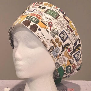 Op de afbeelding: Een witte scrubcap met een kleurrijk patroon van cartoon illustraties en tekst uit de tv-serie "Friends". Het ontwerp bevat het Central Perk logo, voedsel en karakter referenties. De cap heeft een groene band.