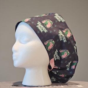 May include: A dark blue surgical scrub hat with a pink tie. The hat has a repeating pattern of green and pink dinosaurs with the text "Thoracic Park" on each dinosaur.