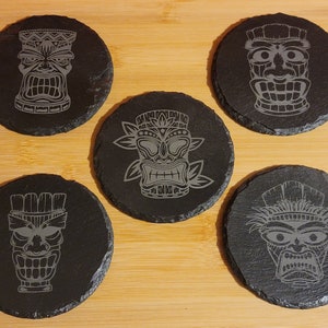 Puede incluir: Conjunto de cinco posavasos de pizarra negra con diseños de máscara tiki grabados. Cada posavasos presenta una máscara tiki diferente con detalles intrincados.