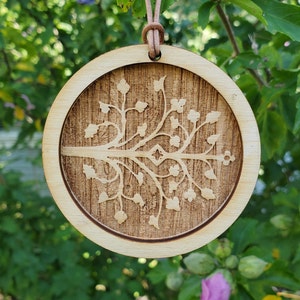 May include: A round wooden ornament featuring a laser-cut design of a tree with branches and leaves. The ornament is suspended from a brown leather cord.