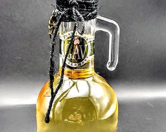 May include: A clear glass bottle with a cork stopper and a black rope handle. The bottle is filled with a light yellow liquid and contains a small leaf. The label on the bottle reads "Gourmet Garlic Oil Vermont".