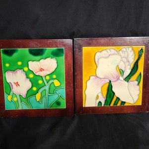 May include: Two framed ceramic tiles with floral designs. The tile on the left has a green background with white and pink flowers. The tile on the right has a yellow background with a white and pink flower.