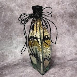 Pode incluir: Uma garrafa de vidro decorativa cheia de vários grãos e sementes, incluindo feijão branco, milho e detalhes em preto. A garrafa é selada com barbante e laço pretos. A garrafa tem um formato alongado único.