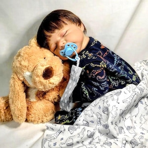 May include: A sleeping baby doll wearing a navy blue onesie with colorful animal designs, a blue pacifier, and a light brown teddy bear. The baby is covered with a white blanket with a dinosaur pattern.