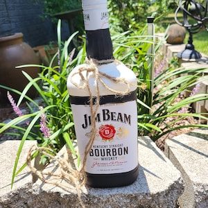 Pode incluir: Uma garrafa de uísque bourbon Jim Beam pintada com um design branco e preto, amarrada com barbante. O rótulo diz "Kentucky Straight Bourbon Whiskey". A garrafa é exibida ao ar livre em uma parede de pedra.