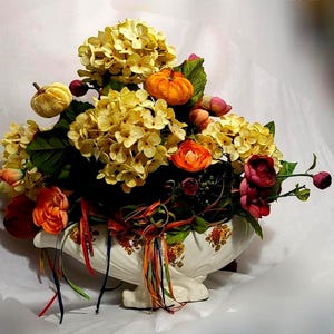 May include: A decorative white ceramic bowl with floral patterns containing an arrangement of yellow hydrangea blossoms, orange pumpkins, and various red and orange flowers, accented with thin, dangling multicoloured ribbons.