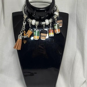 Puede incluir: Un collar plateado con múltiples colgantes, incluyendo tazas de café en miniatura y una borla marrón. Los colgantes de tazas de café presentan varios diseños y colores, con la palabra "COFFEE" visible en algunos. El collar se exhibe en un maniquí negro.