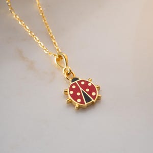 Puede incluir: Un collar dorado con un colgante de mariquita. El colgante de mariquita es rojo con lunares blancos y detalles negros. El collar está sobre una superficie blanca.