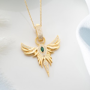 May include: Gold-toned pendant necklace featuring a stylized bird design. The pendant has a central emerald-green stone and is adorned with clear crystals. The necklace hangs on a delicate gold chain, set against a white and light blue background.
