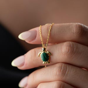 May include: A gold-colored necklace featuring a turtle pendant with a green stone inlay. The turtle pendant is suspended from a delicate chain. The necklace is held in a person's hand, with a black background.