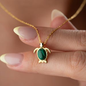 May include: Gold-toned necklace featuring a turtle pendant with a vibrant green stone inlay. The turtle design includes detailed flippers and head, suspended from a delicate chain. The necklace is displayed against a soft, blurred background.