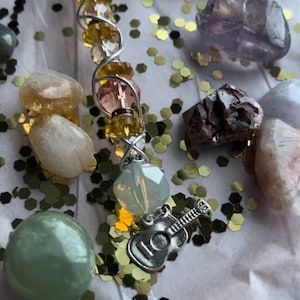 May include: A crystal wand with pink, yellow, and clear beads wrapped around a silver wire. A silver guitar charm hangs from the bottom of the wand. The wand is surrounded by small gold glitter and various crystals.