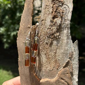 May include: A pair of dangle earrings featuring a vertical silver-toned wire frame containing three rectangular beads. The beads alternate between translucent amber-colored glass and metallic gold-toned segments, suspended from silver-toned ear wires.