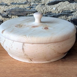 Könnte beinhalten: Eine hellfarbene Holzschale mit Deckel und kleinem Knaufgriff. Schale und Deckel sind rund, mit sichtbarer Holzmaserung und Knoten. Die Schale steht auf einer Holzoberfläche.