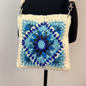 May include: A square crochet crossbody bag featuring a central mandala pattern in shades of light blue, dark blue, and brown. The bag is framed by a cream-colored border and includes a matching cream-colored crochet strap attached with silver metal clips.