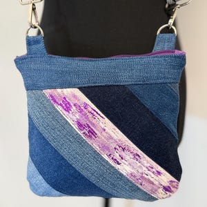 May include: A denim crossbody bag with a purple zipper and strap. The bag features a diagonal stripe of white fabric with purple paint strokes. The bag is made of various shades of blue denim.